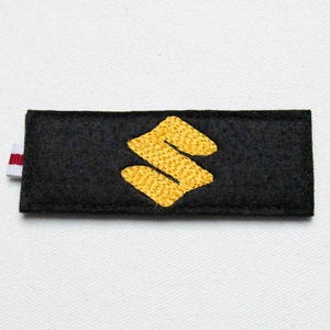 May include: A rectangular black felt patch featuring a gold embroidered Suzuki logo in the center. A small white fabric tab with a thin red stripe is attached to the left side of the patch. The patch has a textured surface and clean edges.
