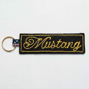May include: A rectangular black fabric keychain featuring the word Mustang embroidered in gold script lettering. The keychain includes a gold metal ring attached by a small fabric tab patterned with the red, white, and blue American flag design.