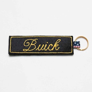 May include: A rectangular black felt keychain featuring the word Buick embroidered in yellow cursive script. The border is stitched in yellow thread, and a small fabric tab with a red, white, and blue star pattern is attached to a gold metal ring.