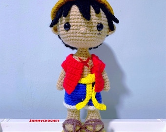 Op de afbeelding: Een gehaakt figuur van een cartoonfiguur met zwart haar, een strohoed, een rood shirt, een blauwe broek en een gele riem. Het figuur staat op een houten voet.
