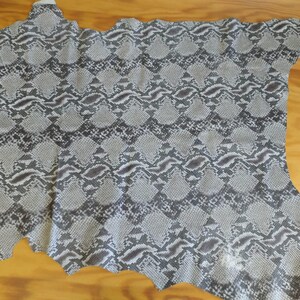 Może przedstawiać: Kawałek skóry węża w kolorze szarym i białym z wzorem diamentowym. Skóra jest miękka i elastyczna, idealnie nadaje się do tworzenia różnych rękodzieł.