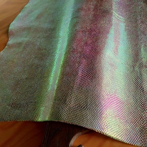 Könnte beinhalten: Schillerndes Lederfell mit schuppenartiger Textur. Das Leder schimmert in Grün-, Lila- und Goldtönen. Das Material ist über eine Holzoberfläche drapiert und zeigt seine Flexibilität und seinen Glanz.