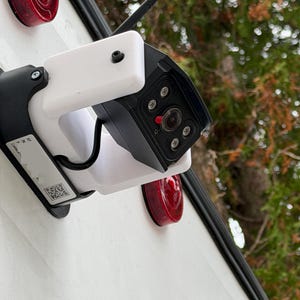 Puede incluir: Una cámara de seguridad en blanco y negro con un soporte de montaje blanco está sujeta a una superficie blanca. La cámara tiene una lente, varias luces pequeñas y una pequeña antena. Luces traseras rojas visibles.