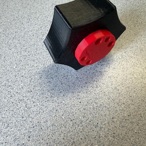 May include: A black, geometric-shaped object with a red, circular component. The black part has a star-like design with pointed edges. The red circle has six holes. The object appears to be 3D-printed.