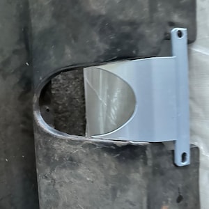 May include: A close-up of a gray metal bracket attached to a black plastic surface. The bracket has a curved shape and is designed to hold something in place.