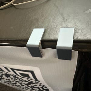 May include: Two silver metal clips are holding a white poster with a black and white QR code printed on it.