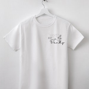 Op de afbeelding: Wit T-shirt met ronde hals, hangend aan een witte hanger. Het shirt heeft een zwarte paardenbloemafbeelding met de woorden "Just Dandy" in cursief. Het ontwerp bevindt zich op de rechterborst.