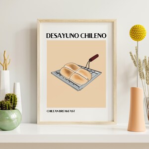 Puede incluir: Un cuadro enmarcado con una ilustración estilizada de un desayuno chileno. La obra muestra cuatro panecillos dorados sobre una parrilla metálica, con los textos "DESAYUNO CHILENO" y "CHILEAN BREAKFAST". El cuadro está en un marco de madera clara.