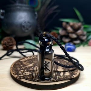 Può includere: Collana con ciondolo a forma di bottiglia di vetro nero e cordoncino nero. La bottiglia è piena di sabbia nera e un simbolo di runa bianco. La bottiglia ha un tappo dorato.