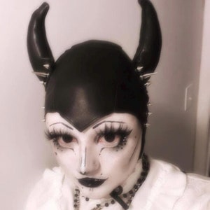 May include: A person wearing a black leather devil horn headpiece with silver spikes, white makeup, and black lipstick. They are wearing a white blouse with a ruffled collar and multiple silver and black necklaces.