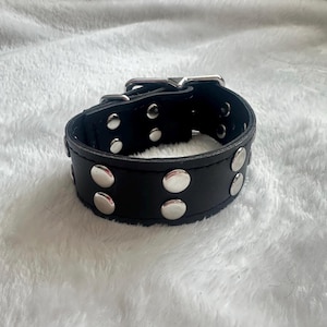 May include: Black leather bracelet with silver-colored studs and a buckle closure. The bracelet is a wide band with multiple rows of studs. The buckle and studs are shiny, contrasting with the matte black leather. A stylish accessory.