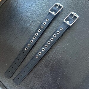 May include: Two black leather straps with silver metal buckles and rows of silver eyelets.