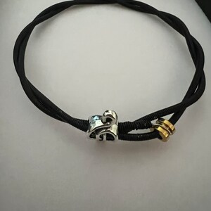 Könnte beinhalten: Ein schwarzes Gummiband mit einem silbernen Anhänger und einem goldenen Anhänger. Der silberne Anhänger ist ein stilisierter Buchstabe 'S' und der goldene Anhänger ist ein einfacher Ring.