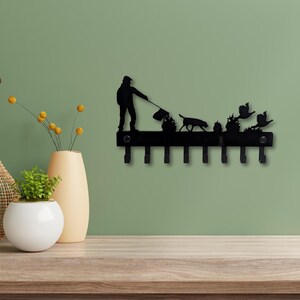May include: Black metal key holder depicting a person walking a dog, with birds and bushes. The key holder has seven hooks. Decorative vases and plants are in the foreground.
