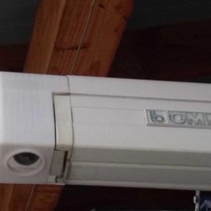 Pode incluir: Um componente de toldo branco com a marca "OMNIS" visível. A estrutura retangular tem uma extremidade arredondada com uma abertura circular. O fundo apresenta vigas de madeira.