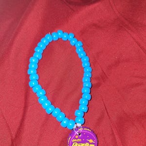May include: A blue beaded necklace with a pendant made from a purple bottle cap. The bottle cap features the text "Nuka Cola Quantum" and "JONES SODA CO." on a purple background with a planet design.