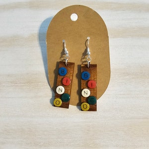 May include: Wooden earrings with the word "BINGO" spelled out with colorful circular accents. The earrings are rectangular and hang from silver hooks. The background is a light wood surface.