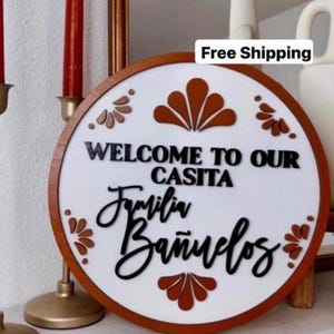 May include: A round wooden sign with a white center and a brown border. The sign reads "WELCOME TO OUR CASITA Familia Bañuelos" in black script. Decorative orange floral accents are placed around the edge.