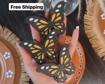 May include: Three wooden butterfly ornaments in black with yellow and white accents. The butterflies are held in a hand.