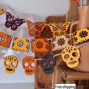 May include: A decorative garland featuring cut-out butterfly and skull shapes in orange, yellow, and black. The garland is strung on twine and is suitable for festive occasions. The background includes a wooden cutting board and a terracotta vase.