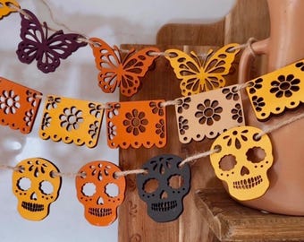 May include: A decorative garland featuring laser-cut wooden shapes strung on twine. The elements include purple and orange butterflies, orange and beige square panels with floral cutouts, and yellow and black sugar skull shapes.