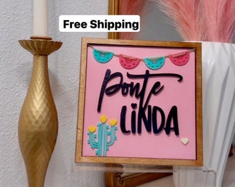 May include: A decorative wooden sign with the words "Ponte LINDA" in black script on a pink background. The sign features a cactus and banner design. A gold candlestick with a white candle is to the left.