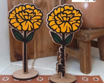 May include: Two wooden marigold flower decorations with yellow petals and black outlines. The flowers have green leaves and black stems. One flower has the name "Maria" cut out. The flowers are on a wooden base.