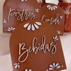 K&ouml;nnte beinhalten: Drei Holzschilder mit gew&ouml;lbten Oberseiten und wei&szlig;en Blumenmustern. Die Schilder sind braun und tragen die Aufschriften "Postres", "Comida" und "Bebidas" in wei&szlig;er Kursivschrift.