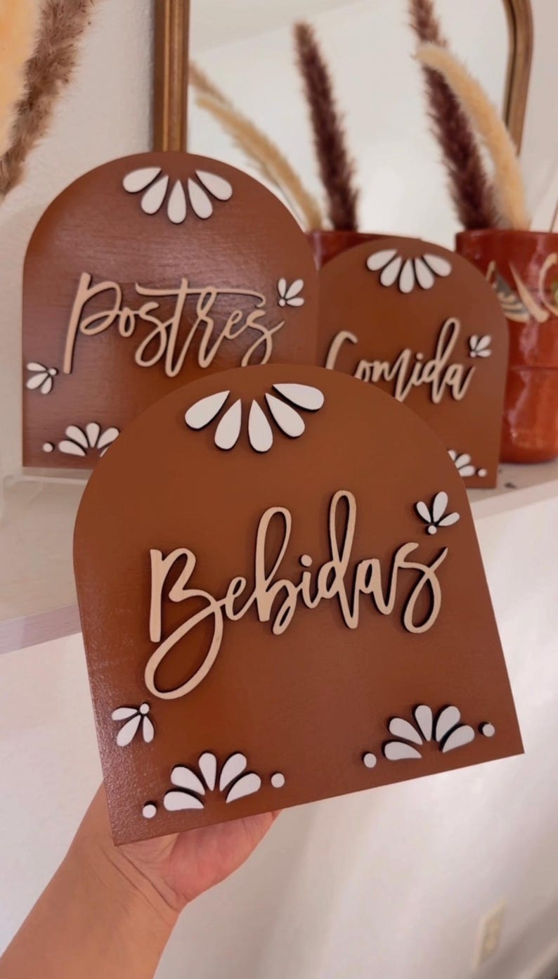 K&ouml;nnte beinhalten: Drei Holzschilder mit gew&ouml;lbten Oberseiten und wei&szlig;en Blumenmustern. Die Schilder sind braun und tragen die Aufschriften "Postres", "Comida" und "Bebidas" in wei&szlig;er Kursivschrift.
