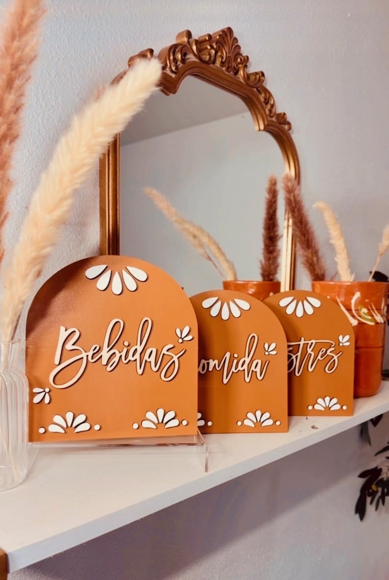 K&ouml;nnte beinhalten: Drei terracottafarbene, gew&ouml;lbte Schilder mit wei&szlig;en Blumenmustern und wei&szlig;en Schriftz&uuml;gen. Die Schilder zeigen "Bebidas", "Comida" und "Stress".