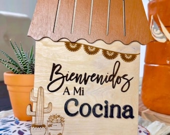 May include: Wooden sign with a house shape and the text "Bienvenidos a mi Cocina" in black lettering. The sign is decorated with a cactus and a small potted plant.