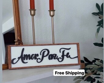 May include: A wooden sign with the words "Amor Paz Fe" written in black cursive lettering. The sign is framed in a dark wood frame and has a white background. The sign is sitting on a shelf with two candlesticks and candles.