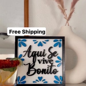 Könnte beinhalten: Ein schwarzes, gerahmtes Holzschild mit weißem Hintergrund und blauen floralen Akzenten. Das Schild trägt die Aufschrift "Aqui Se Vive Bonito" in schwarzen Buchstaben.