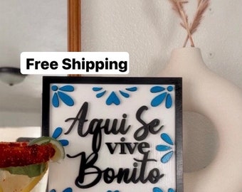May include: A black framed wooden sign with a white background and blue floral accents. The sign says "Aqui Se Vive Bonito" in black lettering.