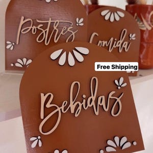 K&ouml;nnte beinhalten: Drei Holzschilder mit abgerundeten Oberseiten und wei&szlig;en Blumenmustern. Die Schilder sind braun und haben die W&ouml;rter "Postres", "Comida" und "Bebidas" in Wei&szlig; geschrieben. Kostenloser Versand steht unten auf dem Schild.