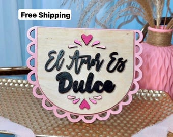 May include: A wooden sign with a pink scalloped border and the text "El Amor Es Dulce" in black lettering. The sign is decorated with pink hearts.