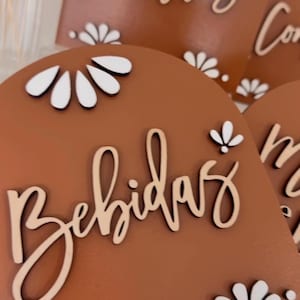 K&ouml;nnte beinhalten: Drei Holzschilder mit wei&szlig;en floralen Akzenten und den W&ouml;rtern "Postres", "Bebidas" und "Comida" in kursivschrift. Die Schilder sind braun und haben einen rustikalen Look.