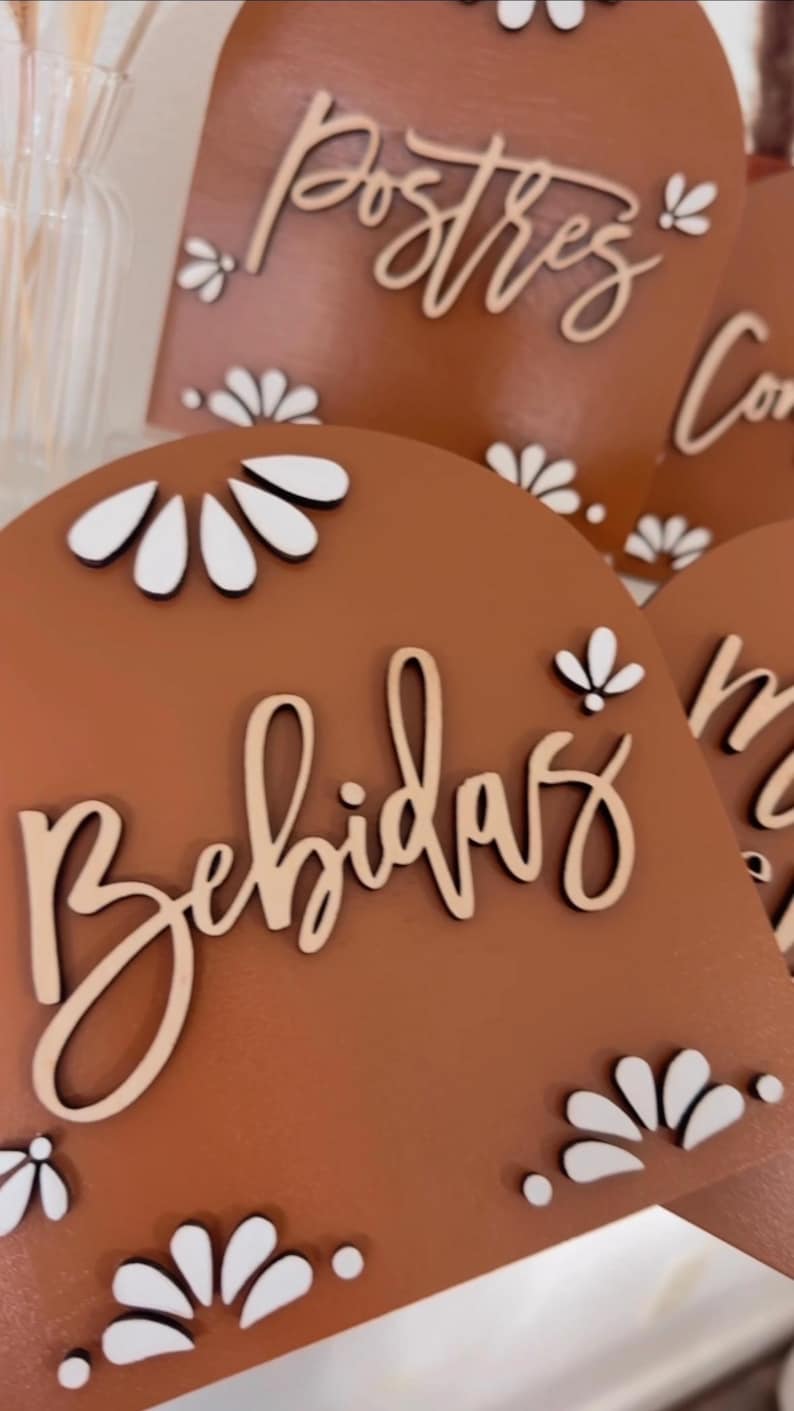 K&ouml;nnte beinhalten: Drei Holzschilder mit wei&szlig;en floralen Akzenten und den W&ouml;rtern "Postres", "Bebidas" und "Comida" in kursivschrift. Die Schilder sind braun und haben einen rustikalen Look.
