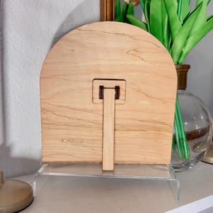 K&ouml;nnte beinhalten: Ein Holzbildrahmen mit hellbrauner Oberfl&auml;che und einem St&auml;nder auf der R&uuml;ckseite. Der Rahmen ist bogenf&ouml;rmig und hat ein schlichtes Design.