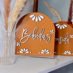 K&ouml;nnte beinhalten: Zwei Holzschilder mit braunem Hintergrund und wei&szlig;en Blumenmustern. Das Schild links sagt "Bebidas" und das Schild rechts sagt "Comida".