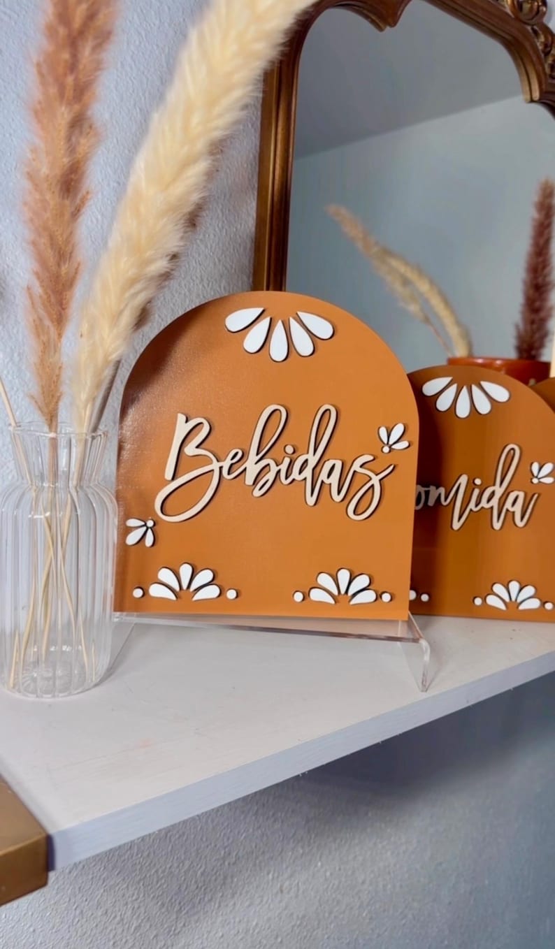 K&ouml;nnte beinhalten: Zwei Holzschilder mit braunem Hintergrund und wei&szlig;en Blumenmustern. Das Schild links sagt "Bebidas" und das Schild rechts sagt "Comida".
