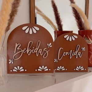 K&ouml;nnte beinhalten: Zwei terracottafarbene Holzschilder mit wei&szlig;en gewellten R&auml;ndern und wei&szlig;en Buchstaben. Das Schild links lautet "Bebidas" und das Schild rechts "Comida".