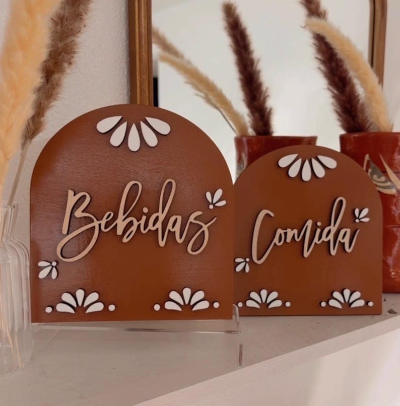 K&ouml;nnte beinhalten: Zwei terracottafarbene Holzschilder mit wei&szlig;en gewellten R&auml;ndern und wei&szlig;en Buchstaben. Das Schild links lautet "Bebidas" und das Schild rechts "Comida".