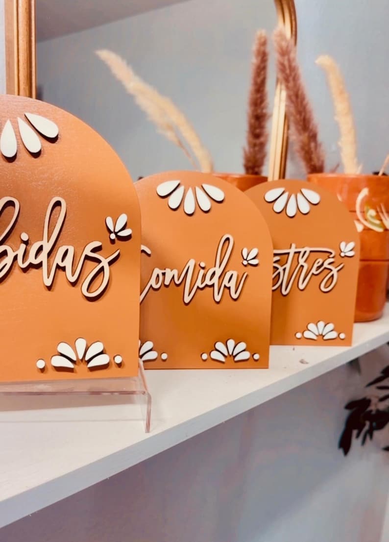 K&ouml;nnte beinhalten: Drei terracottafarbene, bogenf&ouml;rmige Schilder mit wei&szlig;en Blumenmustern und wei&szlig;en Schriftz&uuml;gen. Die Schilder zeigen "Bebidas", "Comida" und "Postres".