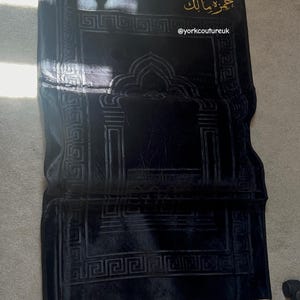 Op de afbeelding: Een zwart gebedskleed met een centraal mihrab-boogontwerp, goudkleurige franjes aan de boven- en onderkant en goudkleurige Arabische kalligrafie bovenaan. De tekst @yorkcoutureuk is in het wit gedrukt nabij de bovenkant van het kleed.
