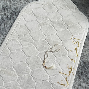 Può includere: Un tappetino da preghiera color crema con un motivo a quadrifoglio in rilievo, frange dorate sul bordo inferiore e calligrafia araba dorata sul lato destro. Un rosario di perline bianche con una piccola nappa poggia sul tappetino.