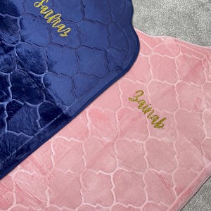 May include: Two rectangular prayer mats with a velvet-like texture and a repeating quatrefoil pattern. One mat is dark blue with the name Zainab embroidered in gold, and the other is light pink with the name Zainab embroidered in gold.