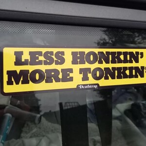 May include: A rectangular yellow sticker with the text "LESS HONKIN' MORE TONKIN'" in bold, dark brown letters. The sticker also includes the word "Deathtrap" in a smaller font. The sticker is affixed to a window.