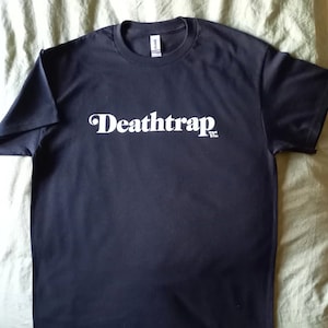 Peut inclure: Un t-shirt noir avec le texte blanc "Deathtrap" imprimé sur le devant.