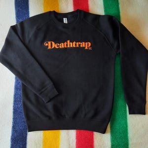 Peut inclure: Sweat-shirt noir avec le texte orange "Deathtrap" sur la poitrine. Le sweat-shirt a des manches longues et un col rond. Le texte est dans une police stylisée. Le sweat-shirt est présenté sur une couverture rayée.